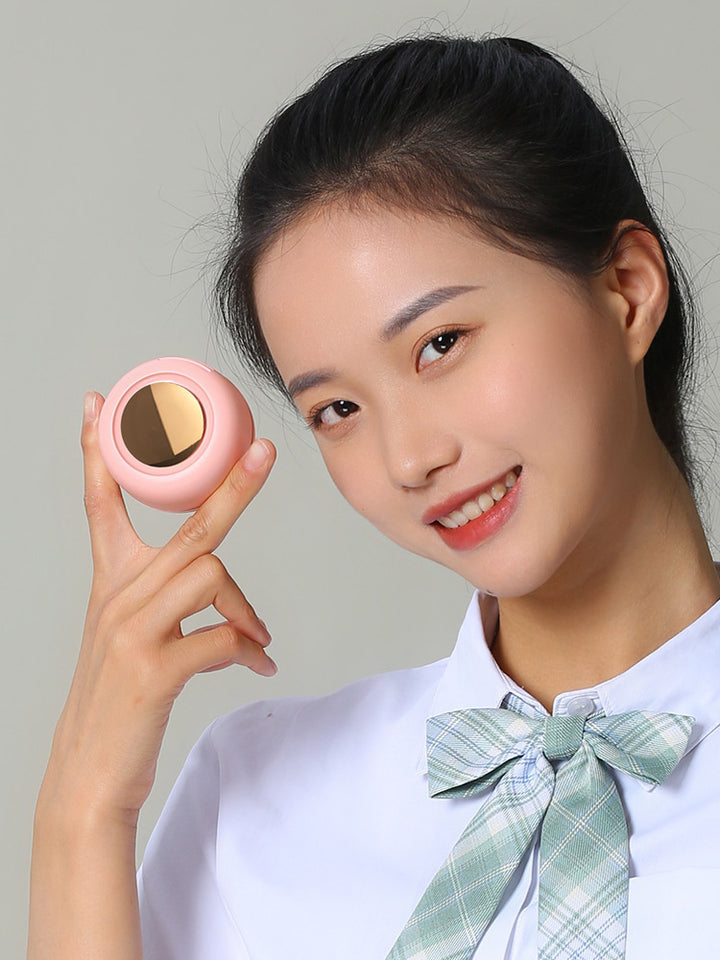 Woman holding a pink compact mirror against a plain background with her mini cooling fan